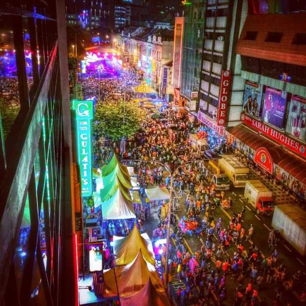 Eine lebhafte Faschings-Szene mit einer großen Menschenmenge auf der Straße, umgeben von Zelten, Geschäften, Gebäuden und Bannern, mit einer hell erleuchteten Fläche im Hintergrund.