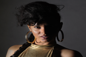 Eine Frau in einem goldenen Kleid und großen Ohrringen steht selbstbewusst vor einer Wand, ihr glatter Bob-Haarschnitt und ihr natürliches Make-up unterstreichen ihren entschlossenen Gesichtsausdruck, während sie direkt in die Kamera schaut.