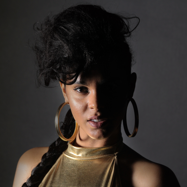 Eine Frau in einem goldenen Kleid und großen Ohrringen steht selbstbewusst vor einer Wand, ihr glatter Bob-Haarschnitt und ihr natürliches Make-up unterstreichen ihren entschlossenen Gesichtsausdruck, während sie direkt in die Kamera schaut.