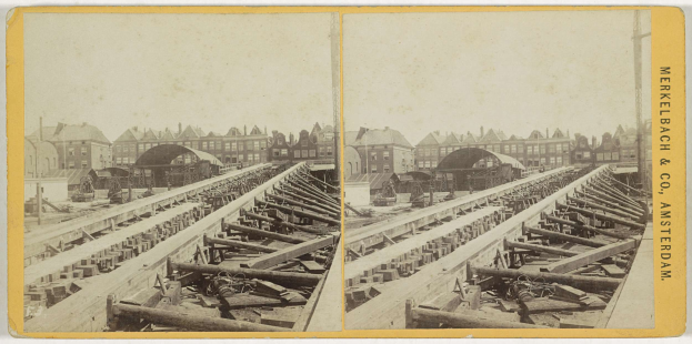 Schwarz-weiß-Foto einer Bahnstrecke mit Gebäuden im Hintergrund, Text 'Merkelbach & Co, Co. Amsterdam' auf der rechten Seite und verstreute hölzerne Gegenstände im Vordergrund.