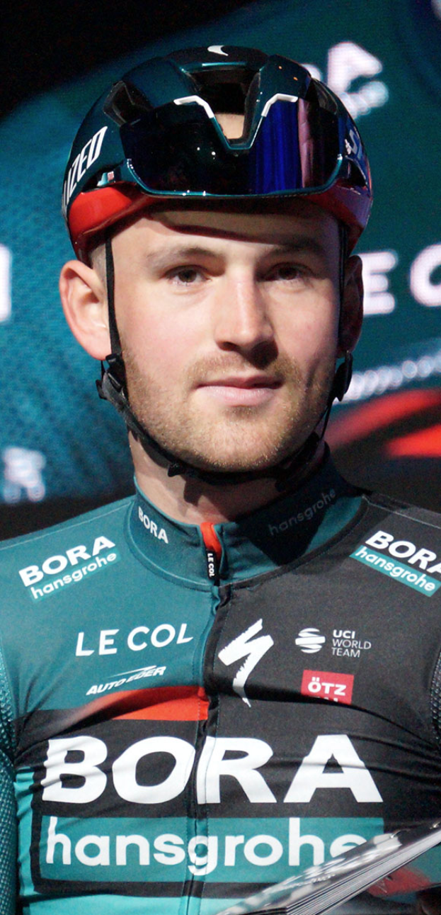 Ein Mann in einem blauen Helm und Sonnenbrille steht neben einem Fahrrad und trägt ein Trikot mit dem Bora-Hansgrohe World Tour-Logo, mit einem entschlossenen Gesichtsausdruck.
