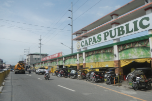 Eine belebte Stadtstraße mit Fahrzeugen, einem Geländer auf der linken Seite, Gebäuden, Pfählen, Laternen, Schildern und einigen Menschen bei bewölktem Himmel, mit einem Gebäude, auf dem ein "Capas Public"-Schild steht.