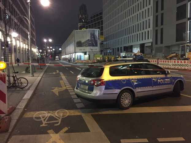 Ein Polizeiwagen parkt nachts auf einer Stadtstraße, umgeben von Gebäuden, Straßenmöbeln, Fahrzeugen und Menschen, mit einer Statue im Hintergrund.