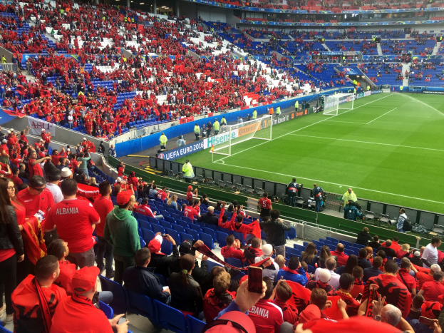 Eine große Menschenmenge in einem Stadion schaut einem Fußballspiel zu, mit Menschen, die sitzen und stehen, Fahnen, einem Torpfosten und Deckenleuchten.