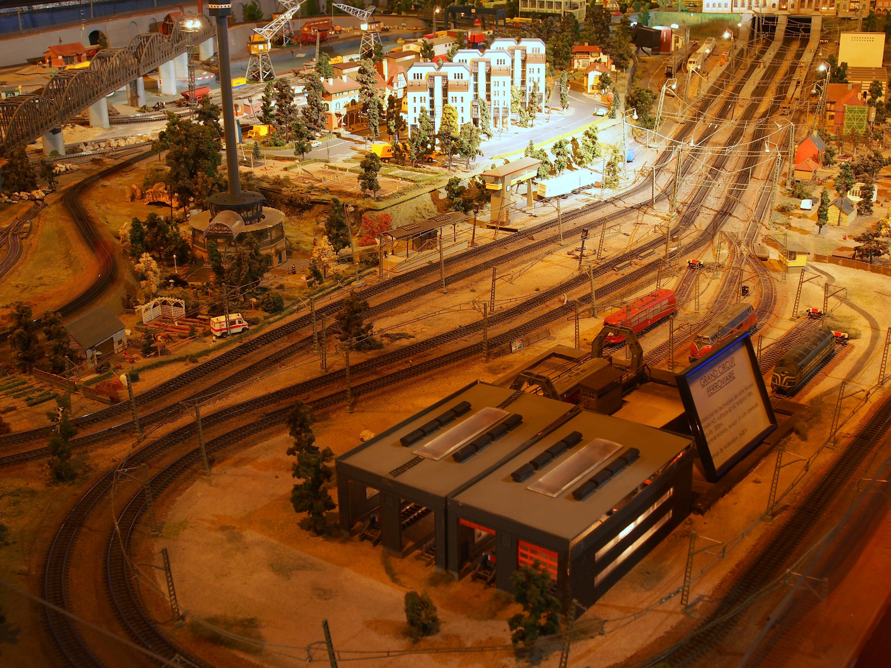 Eine detaillierte Miniatur-Modellbahnanlage in einem Museum, die Gebäude, Bäume, Schienen, Fahrzeuge, Pfosten, Lichter und Platten zeigt, die eine belebte Stadtlandschaft nachahmen.