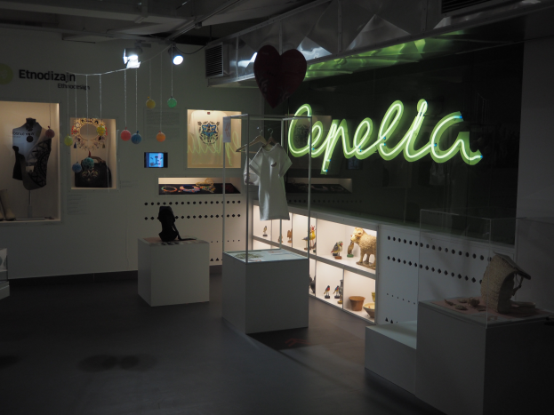 Ein Raum voller verschiedener Gegenstände, darunter Schaufensterpuppen mit Kleidern, Gegenstände auf Regalen, ein Display-Bildschirm, ein Namensschild, dekorative Gegenstände an der Decke und Deckenleuchten bei der Cepella-Ausstellung in Mailand.