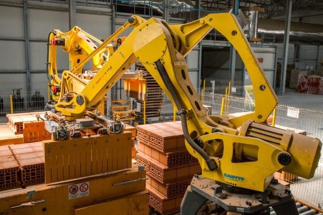 Großes Industriegebäude mit gestapelten Kisten, Kränen, Zaun, Pfählen und einer fernen Wand in einer Fertigungseinrichtung.