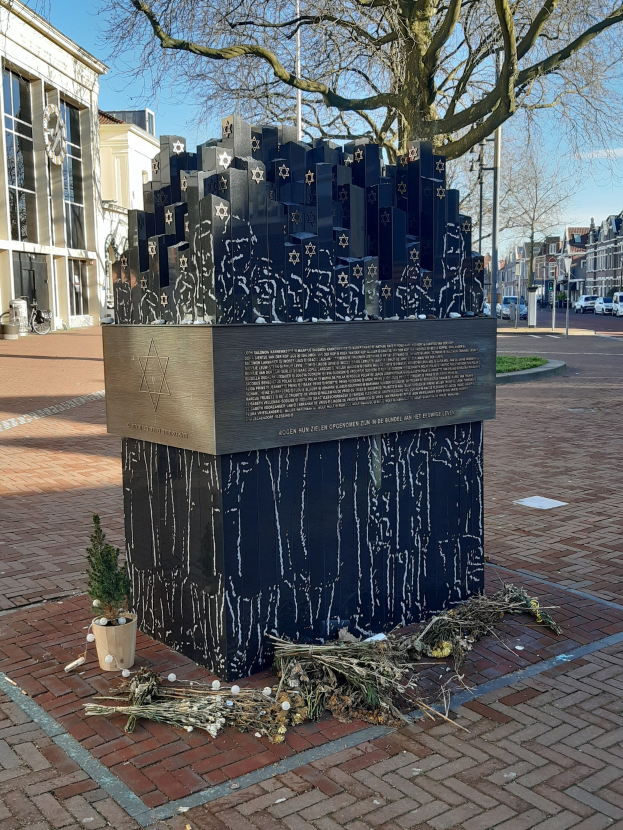 Mahnmal für Holocaust-Opfer in einem Stadtplatz mit einem Baum davor, einem Topfgewächs, verstreuten trockenen Blättern und umliegenden Gebäuden, Fahrzeugen, Pfählen und Bäumen.
