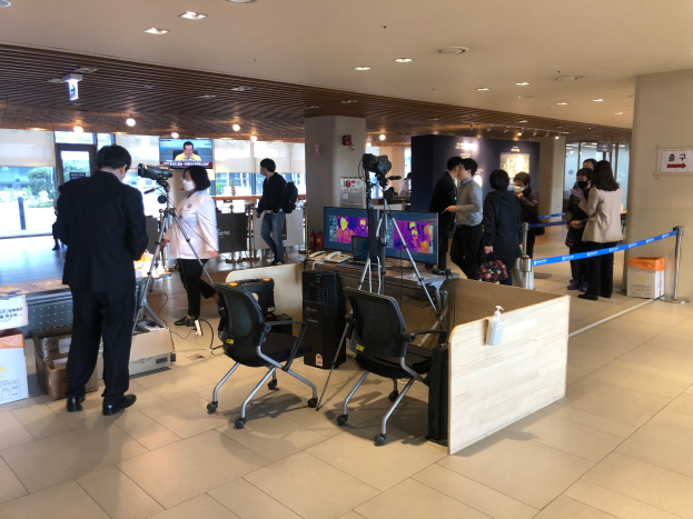 Eine Gruppe maskierter Sicherheitskräfte steht um einen Tresen in einem Flughafen herum, mit Stühlen, Tischen, Monitoren und einer Absperrung mit Bändern in der Nähe, Kartons auf dem Boden und ein Schild und Deckenleuchten im Hintergrund.