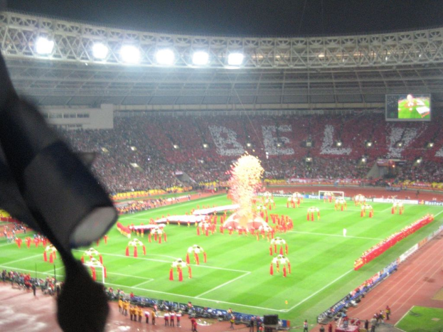 Ein Stadion mit Flutlicht, Eisenstäbe, Zuschauern, stehenden Personen, Werbetafeln, Anzeigetafeln, Überwachungskamera und Fahnen und Dekorationen.