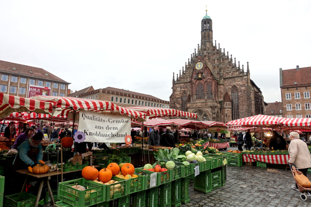 Ein belebter Markt in Nürnberg, Deutschland, mit verschiedenen Früchten und Gemüsen auf dem Stand, Menschen, die umherlaufen, Zelte, Gebäude mit Fenstern und ein Uhrenturm im Hintergrund unter einem sichtbaren Himmel.