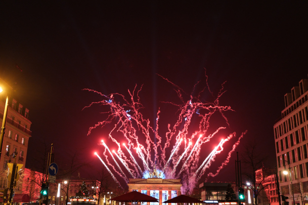 Eine belebte Stadtstraße am Silvesterabend in Berlin, voller Menschen, Fahrzeuge und Gebäude, beleuchtet von festlichen Lichtern und Feuerwerk am Himmel.