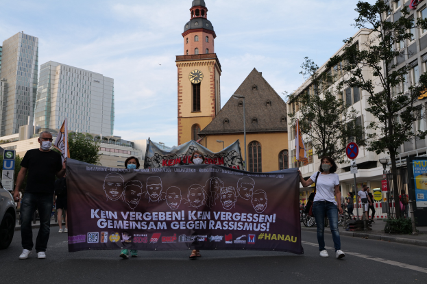 Eine Gruppe von Menschen mit Masken, die eine Fahne mit Text und Bildern tragen, geht eine Straße entlang, mit einem geparkten Auto auf der linken Seite, Gebäuden, Bäumen, Schildern, Pfählen, einem Kirchturm und einem klaren blauen Himmel im Hintergrund.