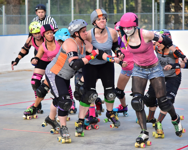 Mehrere Frauen mit Helmen und Schlittschuhen schieben sich gegenseitig auf Kufen, ein Mann steht daneben, ein Metallzaun und Bäume sind im Hintergrund zu sehen.