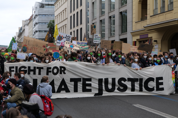 Eine große Gruppe von Menschen marschiert mit einem Banner, auf dem 'Kampf für Hassjustiz' steht, eine Straße entlang, wobei einige Masken tragen und Schilder oder Taschen halten, vor einem Hintergrund aus Gebäuden, Bäumen und einem klaren blauen Himmel.