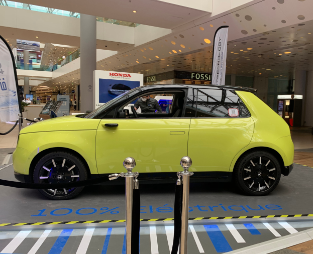 Ein gelbes wasserstoffbetriebenes Elektroauto steht drinnen auf einem Parkplatz, umgeben von Absperrpollern, Bannern und Schildern, mit einer Person darin und einer Gruppe von Menschen in der Nähe.