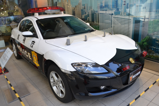 Polizeiauto vor einer Glaswand geparkt, mit einem sichtbaren Rad und einer Texttafel daneben, die Gebäude, Bäume und Himmel durch die Glaswand zeigt.