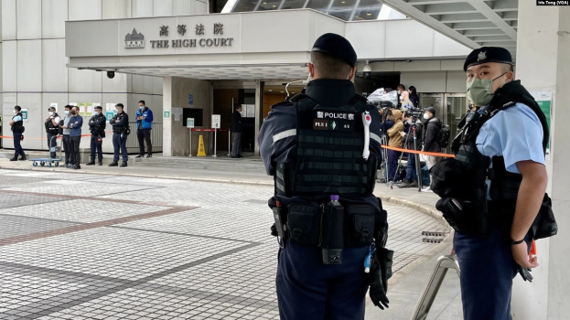 Zwei Polizisten in Uniform, Mützen und Masken stehen vor einem Gebäude am Hong Kong International Airport, umgeben von einer Gruppe von Menschen, einige halten Kameras, mit Pfählen, die mit Bändern geschmückt sind, im Hintergrund.