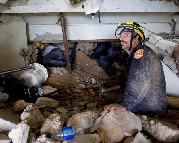 Ein Feuerwehrmann in einem Helm sitzt zwischen Trümmern, darunter Steine und Flaschen, mit einer teilweise sichtbaren Wand mit Text im Hintergrund.