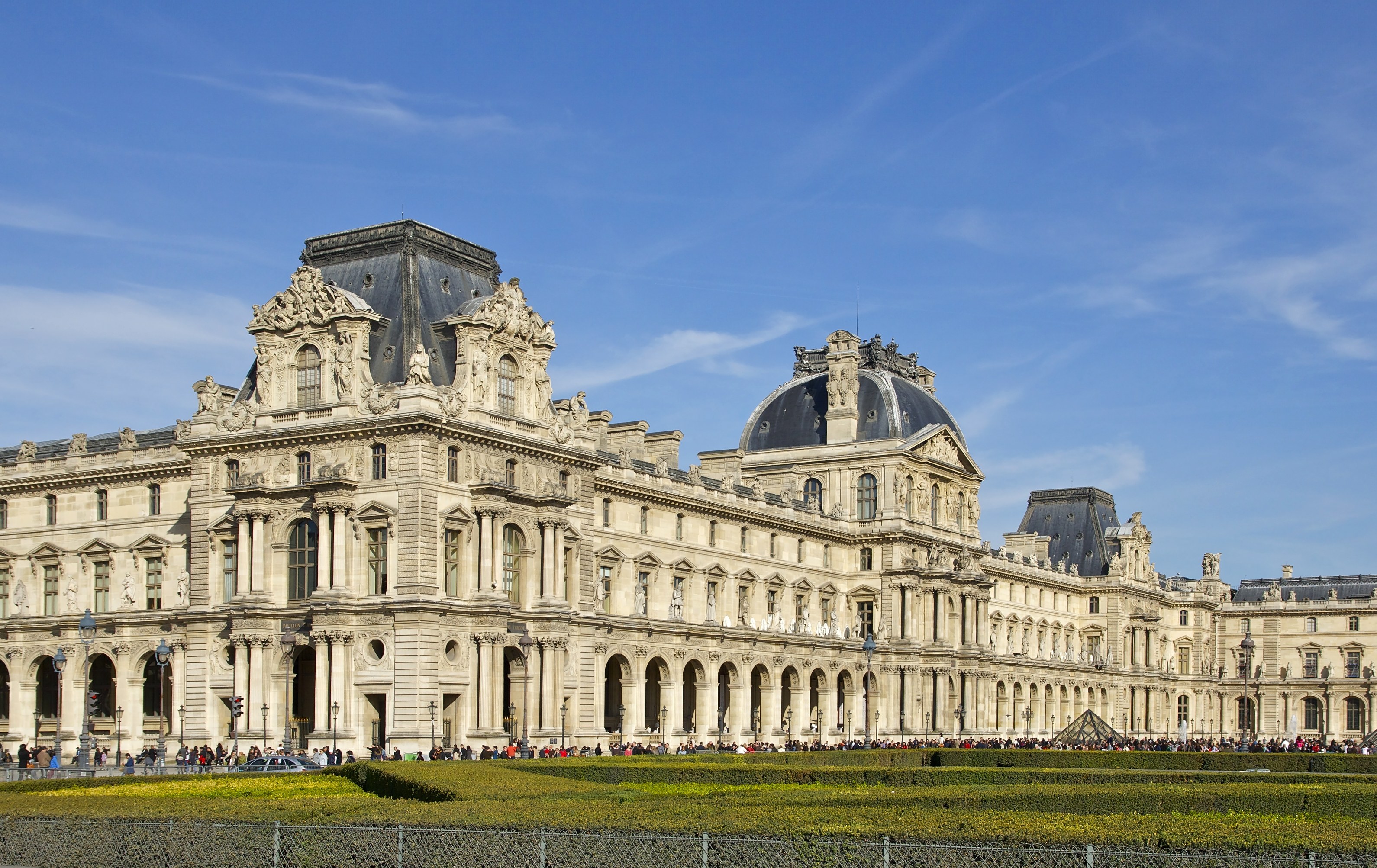 Großes neoklassisches Gebäude mit Säulen und Überdächern, das Louvre-Museum in Paris, umgeben von Menschen, Grün und urbanen Elementen unter einem bewölkten Himmel.