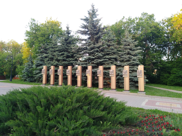 Eine Parkszene mit einem Gedenktafel für Opfer des Tschernobyl-Unfalls, umgeben von Bäumen, Blumen, Gras, einem Weg und einem Straßenmast bei bewölktem Himmel.