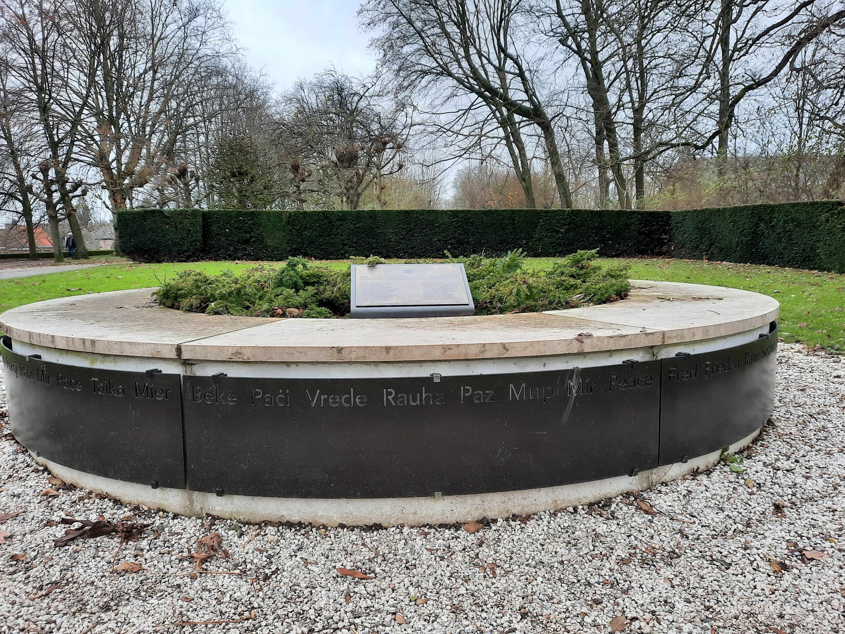 Denkmal mit einer Plakette in einem Park umgeben von Gras, Bäumen, Häusern und einem bewölkten Himmel.