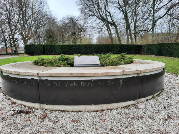 Denkmal mit einer Plakette in einem Park umgeben von Gras, Bäumen, Häusern und einem bewölkten Himmel.