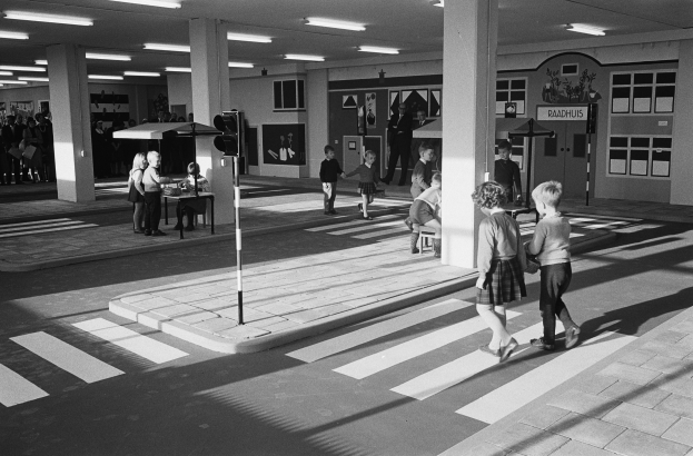 Schwarzes und weißes Foto von Kindern, die eine Straße an einer Fußgängerampel überqueren, mit Fußgängern in der Nähe, einem Verkehrssignalmast im Vordergrund und Architekturpfeilern mit Deckenleuchten im Hintergrund.