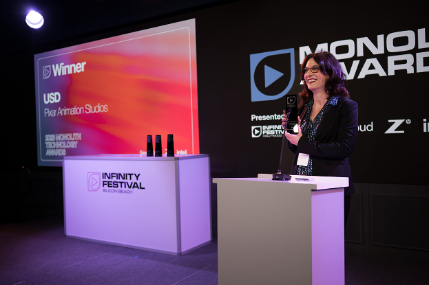 Eine Frau in einem schwarzen Kleid steht an einem Rednerpult und hält eine Trophäe in der Hand, mit einem Mikrofon und anderen Gegenständen auf dem Pult und einem Werbeplakat und einer Lampe im Hintergrund.