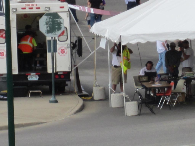 Ein Rettungswagen steht links von einer Stra├če, waehrend auf der rechten Seite ein Zelt eine Gruppe von Menschen sch├╝tzt, mit einem Tisch und einem Stuhl in der N├Ąhe.