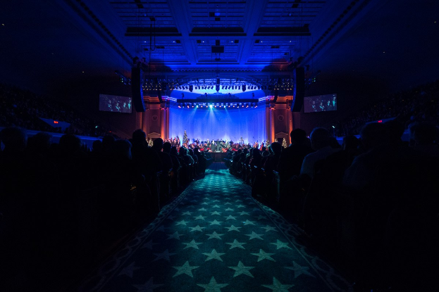 Ein großer Saal mit Stühlen, eine Bühne mit Musikern und eine Decke mit Lampen und Fernsehern, was auf eine Konzerthalle für die Nationale Convention 2019 hindeutet.