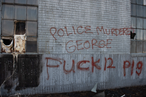 Ein Gebäude mit Graffiti, das "Polizei Mord George Fuck 12 P19" auf der Seite trägt und Fenster sowie verschiedene Gegenstände vorne hat.
