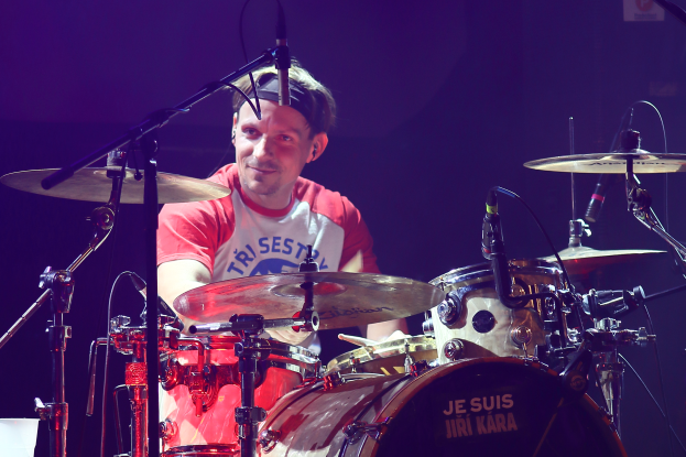 Ein Mann spielt Trommel auf der Bühne bei einem Konzert, trägt ein rotes und weißes T-Shirt und lächelt, mit mehreren Mikrofonen vor ihm.