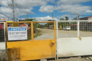 Ein gelbes Tor mit einem Sicherheitshinweisschild, dahinter Strommasten, Bäume, Schuppen, Fahrzeuge und ein bewölkter Himmel.