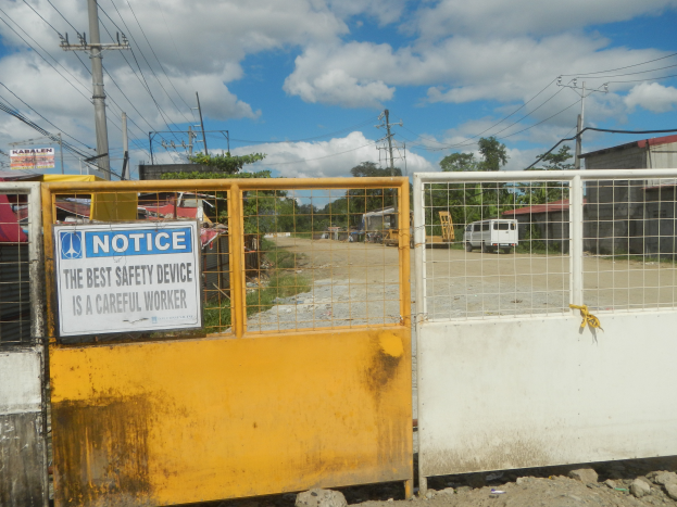 Ein gelbes Tor mit einem Sicherheitshinweisschild, dahinter Strommasten, Bäume, Schuppen, Fahrzeuge und ein bewölkter Himmel.