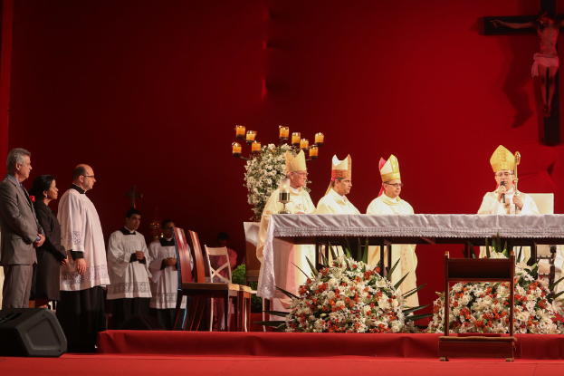 Vier Personen in einem Raum mit Kerzen auf Tischen, eine Person mit Mikrofon, Stühle, Lautsprecher, Blumen und Kreuze an der Wand im Hintergrund.