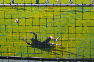 Ein Fußballspieler taucht ab, um einen Ball vor einem Tor zu halten, mit einem Netz im Vordergrund und einigen Menschen im Hintergrund, während mehrere Fußballschuhe auf dem Boden verstreut sind.