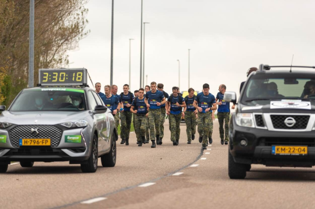 Gruppe von Menschen, die auf einer Straße neben geparkten Autos gehen, mit Bäumen auf der linken Seite und Masten im Hintergrund, unter einem sichtbaren Himmel, an einer Europäischen Cross-Country-Challenge teilnehmend.