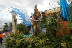 Eine Gruppe von Menschen geht eine Straße entlang neben einem Karnevalswagen, der mit Pflanzen und Blumen geschmückt ist, mit kostümierten Personen auf dem Wagen, Gebäuden, Bäumen und Strommasten mit Drähten im Hintergrund unter einem bewölkten Himmel.