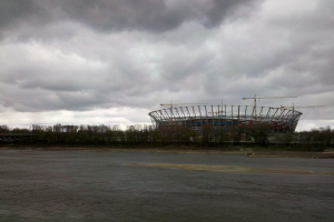 Das Olympiastadion in Kiew, Ukraine, ein großes Stadion umgeben von Bäumen und einer Brücke, mit einem bewölkten Himmel im Hintergrund und sichtbarem Boden unten.