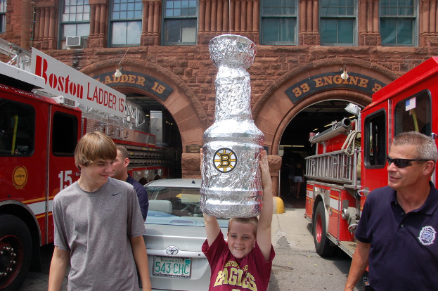 Ein Kind in der Mitte hält eine große flaschenförmige Statue, flankiert von zwei Feuerwehren, einem Auto dahinter und einem großen Gebäude im Hintergrund.