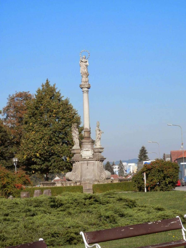 Ein Denkmal in einem Park mit zwei Bänken darauf, umgeben von Pflanzen, Gras, Bäumen, Straßenlaternen, einem Auto auf der Straße, Gebäuden und einem bewölkten Himmel.