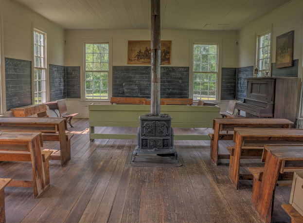 Leerer Klassenraum mit hölzernen Tischen und Stühlen in Reihen, einem zentralen Pfosten, einem Piano auf der rechten Seite und Wänden mit Fenstern, Tafeln und einer Aussicht auf Bäume draußen.