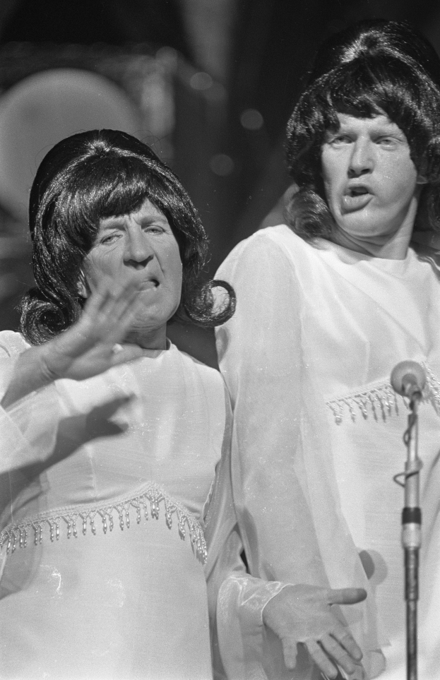 Zwei Frauen in weißen Kleidern stehen Seite an Seite auf einer Bühne, mit einem Mikrofon davor, und scheinen zu singen, auf einem Schwarz-Weiß-Bild.