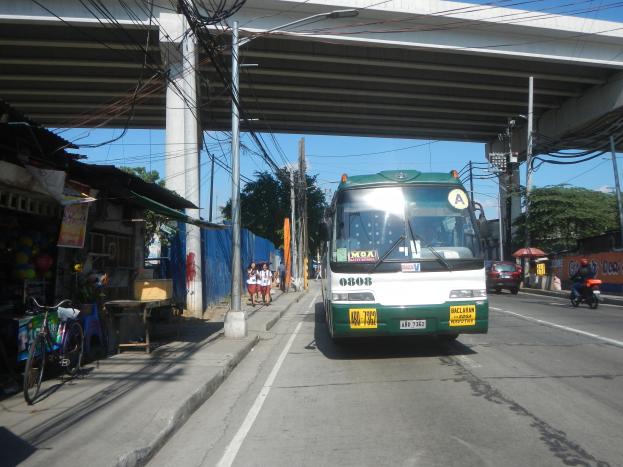 Ein grüner und weißer Bus fährt eine von Geschäften und Ständen gesäumte Straße entlang, unterquert eine Brücke mit Strommasten und -leitungen und fährt an Passanten vorbei, die auf dem Gehweg gehen und Fahrräder in der Nähe abgestellt haben, während im Hintergrund Bäume und ein klarer Himmel zu sehen sind.