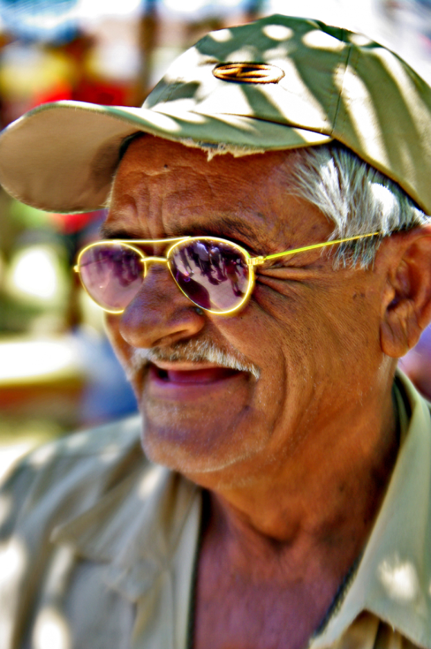 Ein alter Mann mit Mütze und Sonnenbrille, der lächelt, vor einem unscharfen Hintergrund.